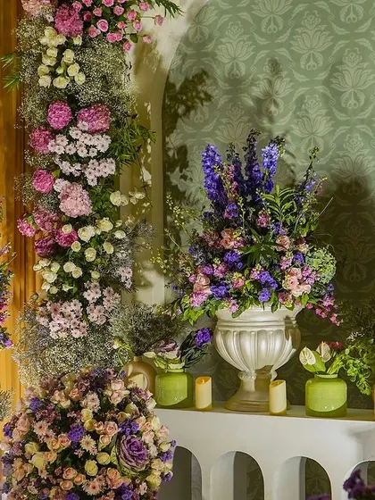 A beautiful vignette featuring large arrangements of lilac, pink, and purple flowers in classic white urns. The arrangements are set within a white arch, creating a stunning floral frame.