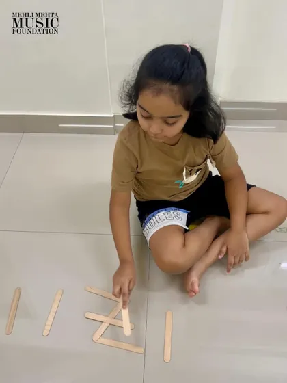 Another young learner concentrates on arranging popsicle sticks to represent a rhythm pattern. We encourage creative, tactile methods to reinforce musical learning.
