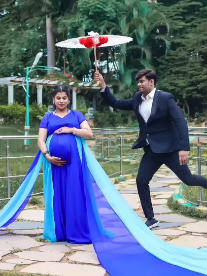 A playful maternity photo of a couple in a park. The father-to-be leaps in the air with an umbrella while his partner looks on in her beautiful blue trail gown.