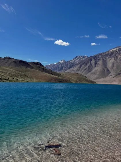 The crystal-clear, turquoise water of Chandratal Lake on a perfect sunny day. You can see the rocks at the bottom near the shore. It's a paradise for photographers and nature lovers.