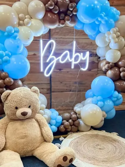 A circular balloon frame with a 'Baby' neon sign makes for a perfect photo booth, featuring a large, cuddly teddy bear.