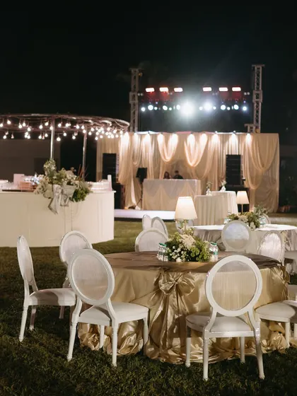 An elegant dinner setup for an evening soirée in Goa. The round tables are draped in gold fabric, with classic white chairs and soft lamp lighting creating a warm and inviting atmosphere.