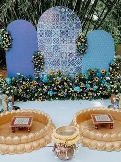 A unique haldi or mehendi setup with a Moroccan-inspired tile backdrop. The blue and white patterns are complemented by sunflowers and blue florals, and the lotus-shaped brass urlis for seating add a distinctive and elegant touch.
