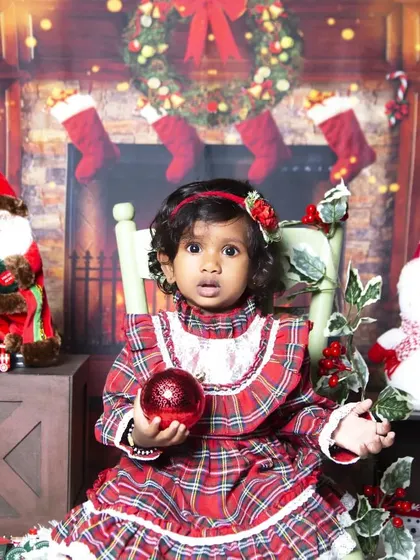 A classic Christmas portrait in front of a cozy fireplace. The toddler is dressed in a festive plaid dress, holding a shiny red ornament.