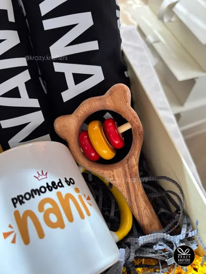 A close-up of the items in our 'Nana-Nani' announcement box, showing the personalized mug and a classic wooden baby rattle.