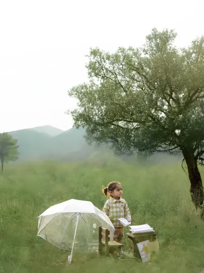 A little girl stands in a misty green field with a miniature desk and a clear umbrella. This imaginative and whimsical setup turns the natural landscape into a storybook scene, perfect for a creative pre-birthday shoot.