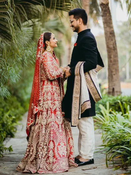 A full-length portrait of the couple, showcasing their stunning red and black wedding attire. The way they hold hands and look at each other creates a classic, romantic image.