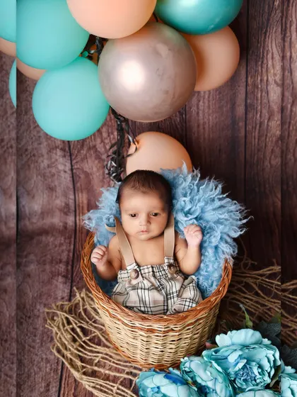 A cute baby boy in plaid overalls sits in a basket, surrounded by a hot-air balloon themed setup with blue and peach balloons.