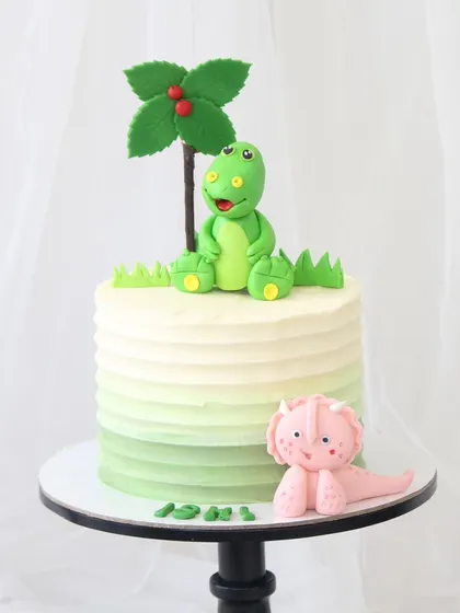 A cute dinosaur-themed cake with an ombre green buttercream finish. The cake is topped with a friendly green dinosaur under a palm tree, with a little pink dino friend at the base.