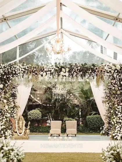 A beautiful wedding stage set inside a transparent tent, allowing for an outdoor feel with indoor comfort. The stage is heavily decorated with white flowers and features a Ganesha idol for blessings.