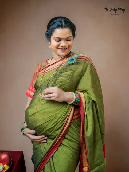 A close-up shot focusing on the mother-to-be's joyful expression and the details of her traditional attire. The peacock feather gently brushing her baby bump adds a symbolic and tender touch.