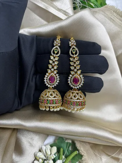 A pair of traditional gold-plated jhumka earrings. These feature intricate work with AD stones and multi-colored accents, making them a timeless and essential item for any ethnic wear collection.