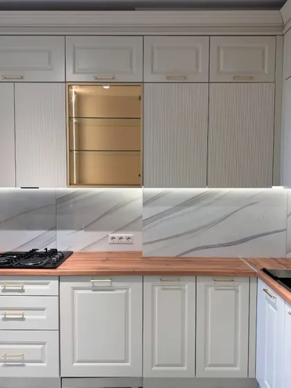 A classic white kitchen with a warm wooden countertop. The design features elegant moulded doors and a special crockery unit with a gold-finished interior and glass shelves for a beautiful display.