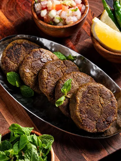 A serving of Shami kebabs with mint leaves. The dark plate and wooden background create a rustic and earthy feel.