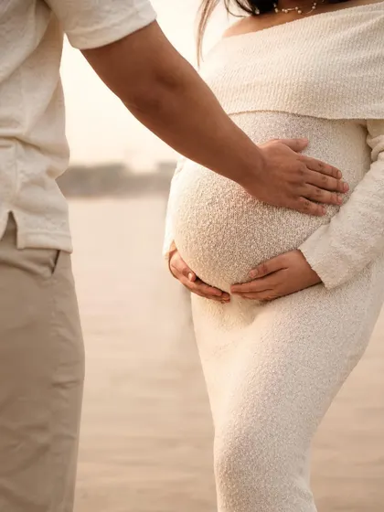 A close-up shot focusing on the hands of both parents cradling the baby bump, with the ocean in the background.