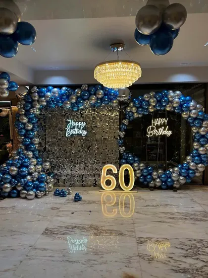 A sophisticated setup for a 60th birthday celebration. This design features two 'Happy Birthday' neon signs against silver sequin walls, framed by a large arrangement of blue and silver chrome balloons.