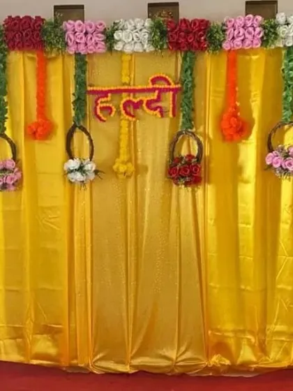 A simple Haldi backdrop using a yellow drape and hanging floral baskets. The 'Haldi' sign is made from red flowers.