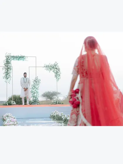 The bride's walk towards the groom. This artistic shot focuses on the groom waiting at the mandap, with the bride approaching in the foreground, creating a sense of anticipation.