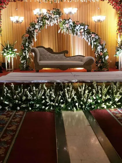A sophisticated stage design with a floral arch in pastel shades. The setup includes a classic diwan sofa, elegant chandeliers, and a fully decorated stage front with white flowers.