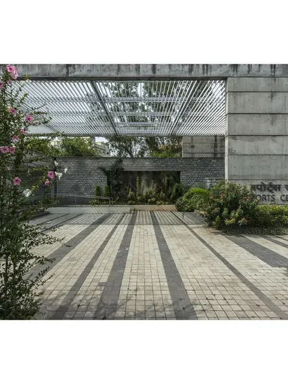 The entrance to the IIM Sports Center, framed by a pergola that casts linear shadows on the ground. The use of stone and concrete unifies the new building with the existing campus.