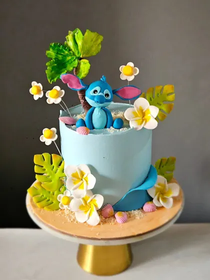 A tropical-themed cake featuring a handcrafted Stitch figurine. The design includes edible flowers and palm leaves, creating a fun, beachy vibe perfect for a Lilo & Stitch fan.