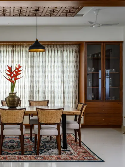 The 'Vasudhalayah' dining area is anchored by a live-edge table and an intricately carved wooden ceiling panel. A large window with bay seating offers a cozy perch for gatherings, flanked by two custom crockery units for elegant storage.
