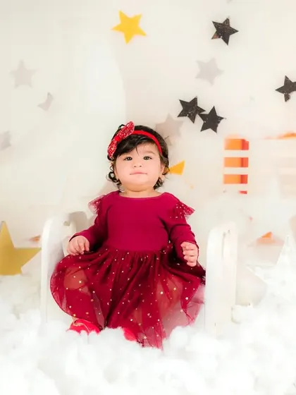 A baby girl in a beautiful red dress sits among star and cloud props, looking serene and dreamy.