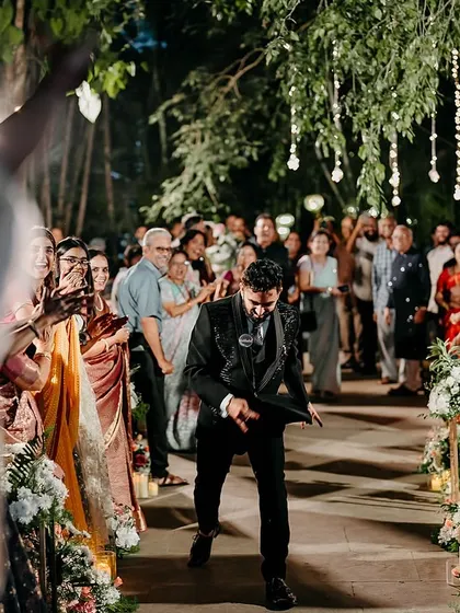 The groom makes a joyful entrance down a beautifully lit aisle, lined with flowers and cheered on by guests.