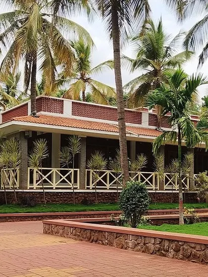 A full view of the 'Grid in the Grove' farmhouse in Sandur. The 8000 sq. ft. structure is seamlessly integrated into the landscape, with coconut trees gracefully adorning the veranda and courtyard.
