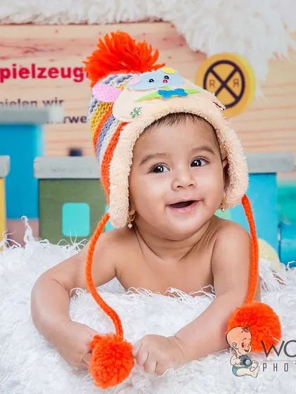 A close-up of a happy baby wearing a colorful winter hat, lying on a soft white blanket with a toy train in the background. His bright smile is sure to warm any heart.