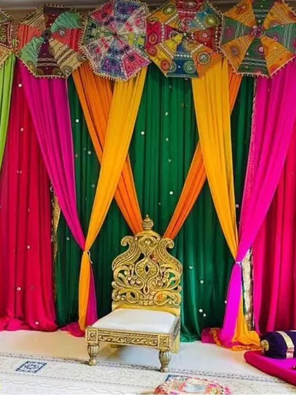 A bright and cheerful Mehendi backdrop with multi-colored drapes in green, pink, and yellow, accented with decorative umbrellas. A single ornate chair is placed for the bride-to-be.