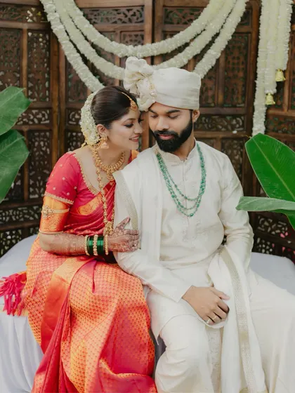 An elegant portrait of the couple seated, the bride in her rich red silk saree and the groom in a classic white sherwani.