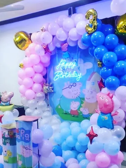 A cheerful Peppa Pig themed birthday party. The decoration features character cutouts against a themed backdrop, surrounded by a colorful balloon arch in shades of pink and blue.