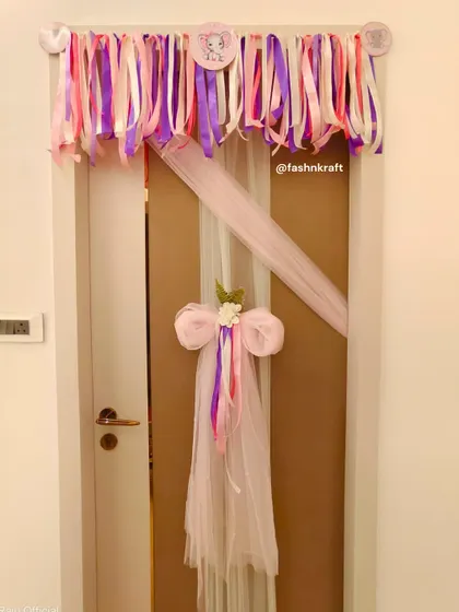 A playful and pretty decoration for a baby girl's room door. A colorful ribbon garland hangs above, while a large tulle bow with trailing ribbons adds a touch of whimsy and charm.
