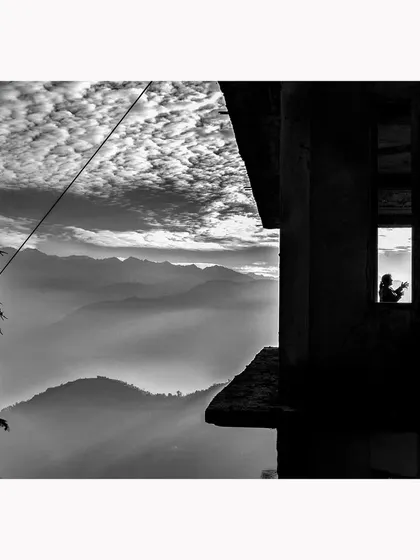 This image frames a quiet, domestic moment within the vastness of a mountain landscape. The silhouette of the figures in the window against the dramatic sky and layered hills creates a story of human life amidst nature's grandeur.