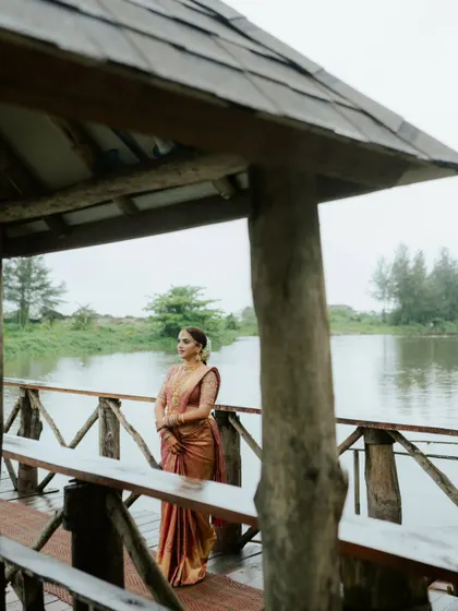 A serene portrait of a South Indian bride in her beautiful silk saree, standing on a wooden pier by the water. The natural setting complements her timeless elegance.