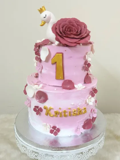 An elegant two-tier cake for a first birthday, featuring a graceful swan topper with a crown, a large mauve sugar rose, and delicate petals.