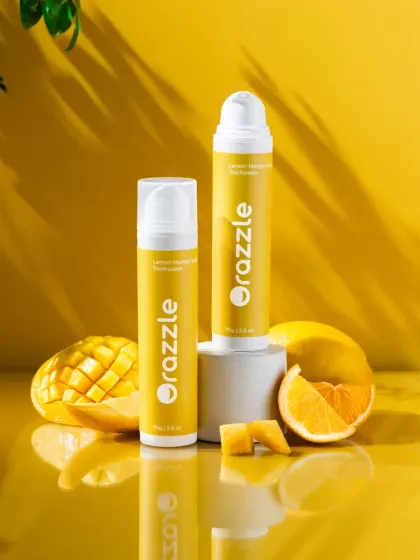 A sunny and vibrant hero shot for Orazzle's lemon-mango toothpaste. The composition uses a glossy, reflective surface and fresh fruit to create a bright, energetic, and high-end look.