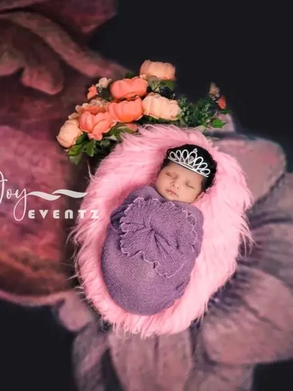A princess-themed newborn portrait where the baby, wearing a tiny tiara, is swaddled in purple and rests on a soft pink fur cushion. The floral background adds to the royal and delicate feel of the photograph.