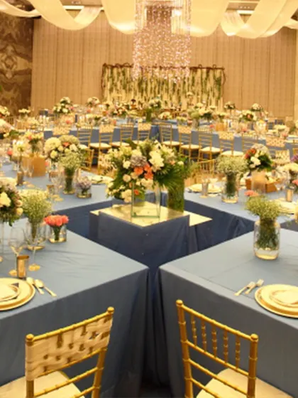 A unique reception layout with tables arranged in a square formation, creating a central space for performances or mingling. The deep blue linens paired with gold chairs offer a sophisticated and modern color palette for an evening celebration.