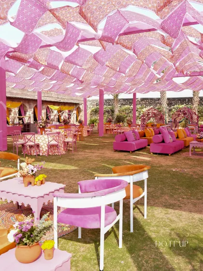 A wide view of the vibrant Haldi seating in Khimsar. The layout includes a mix of sofas and chairs in bright pinks and oranges, with a canopy fatores of block-printed fabric.