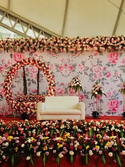 This is a lavish floral stage created for a large reception. The backdrop features a floral print wall, a full floral ring, and a stage lined with a dense arrangement of multi-colored flowers.
