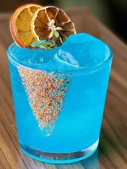 A striking Blue Lagoon cocktail. The vibrant blue color, combined with dehydrated orange slices and a spiced rim, makes for a very Instagrammable drink.