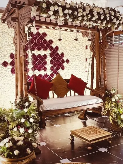 A classic and ornate wooden jhoola (swing) set against a dense wall of white and burgundy flowers. This traditional seating arrangement is perfect for Mehendi or Haldi ceremonies, offering a touch of royal elegance.