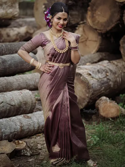 A full-length portrait of the bride in a gorgeous purple silk saree, posing confidently against a rustic backdrop of stacked logs.