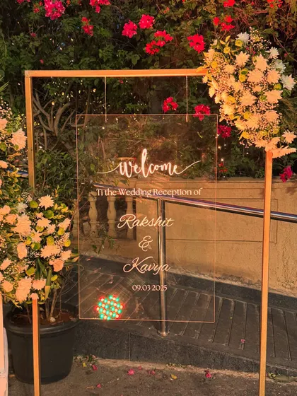 A chic and modern welcome sign for a wedding reception. The clear acrylic board with elegant white calligraphy is framed by a simple gold stand and soft floral arrangements, perfect for a contemporary event.