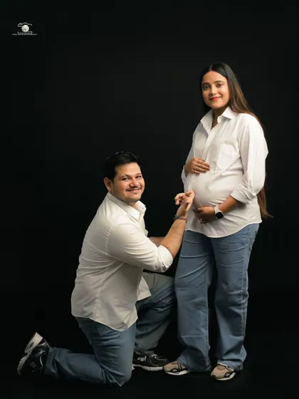 A classic pose with a twist. The dad-to-be kneels and kisses his wife's belly, while she stands smiling in a casual white shirt and jeans against a simple black background.