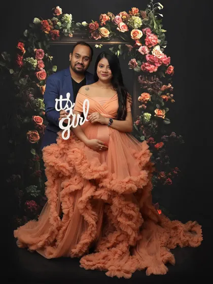 A joyful gender reveal moment captured within a maternity shoot. The couple is framed by a beautiful floral arch, and the mom-to-be wears a stunning ruffled peach gown, adding to the celebratory feel.