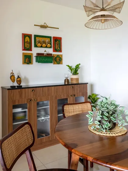A wider view of the puja and crockery area, showing how it integrates with the dining space. The round wooden dining table and cane chairs complement the traditional aesthetic.
