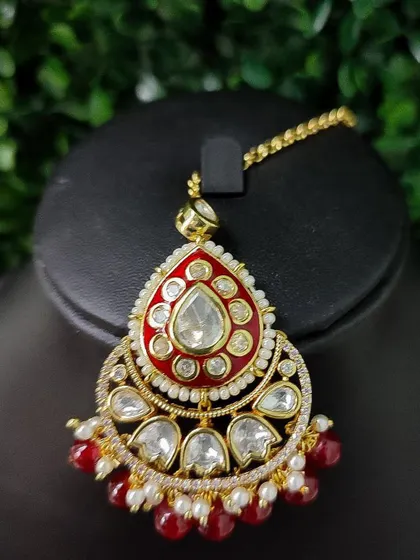 A bold maang tikka with bright red enamel work and kundan stones. The red bead drops complete this eye-catching piece.
